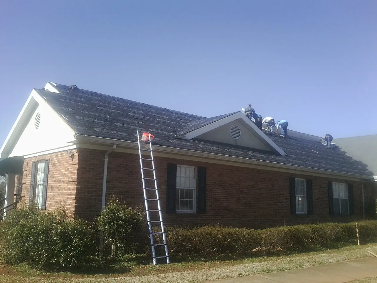 Crew tearing shingles off a brick ranch roof with a ladder set — Emergency Roof Repair work by our crew in Maple Hill