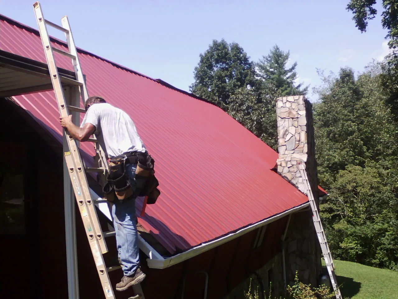 Roofer on a ladder beside a red metal roof and stone chimney — Soffit & Fascia Repair by Roofing Brothers in Maple Hill